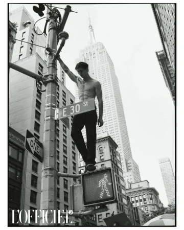 Black and white portrait of a shirtless man standing on a traffic light with the Empire State Building in the background in New York City.