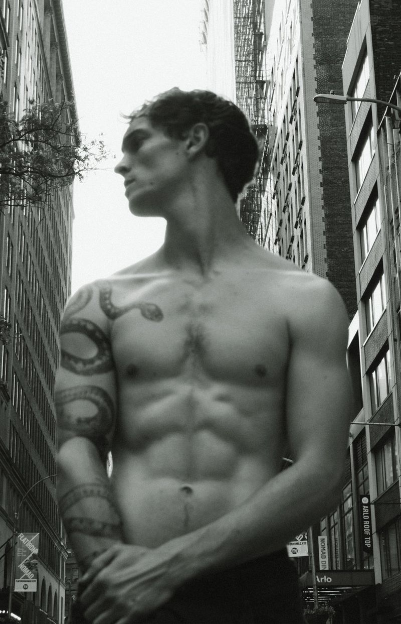 Black and white shot of a muscular, shirtless adult male with a snake tattoo, standing outdoors in an urban environment between tall buildings.