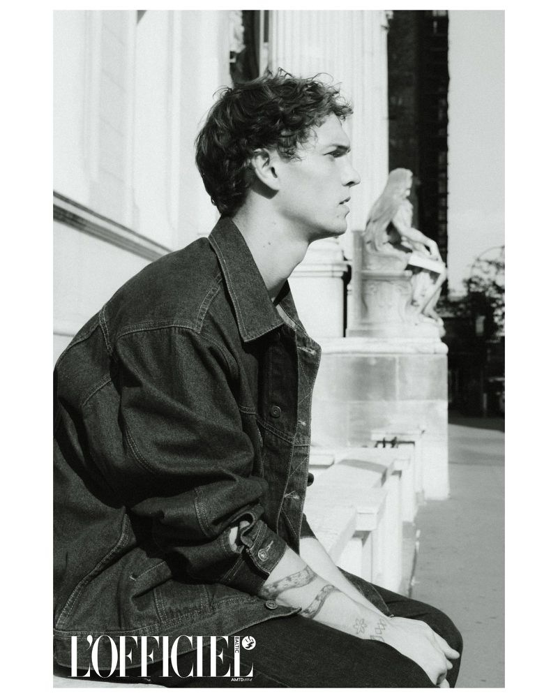 Profile portrait of a young adult man in a denim jacket sitting outdoors on a sunny day near an ornate building with a statue.