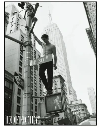 Shirtless male model standing on a city street sign post with skyscrapers in the background.
