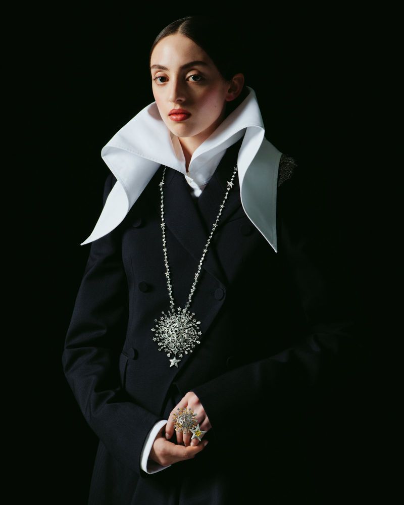 Elegant woman in a black suit with a large white collar, adorned with a star-themed necklace and ring, posing against a black background.