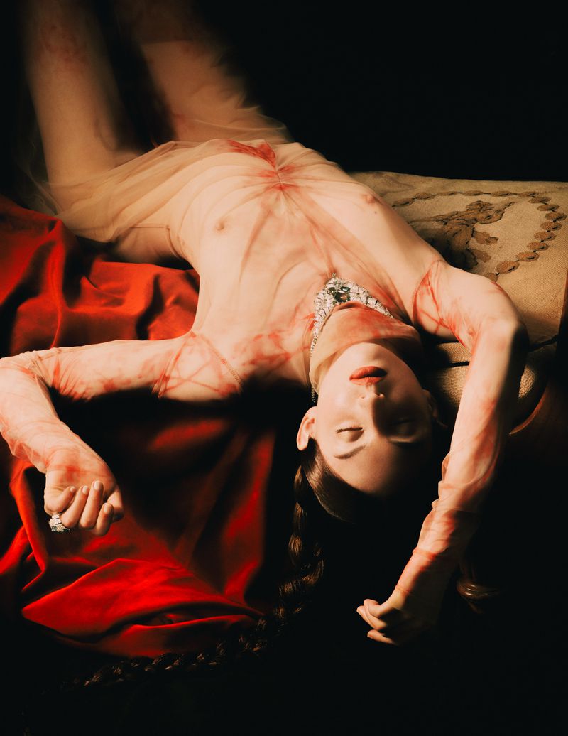 Dramatic fashion photo of a fair-skinned adult female lying down on a red velvet surface, wearing sheer clothing and jewelry in a dark indoor setting.