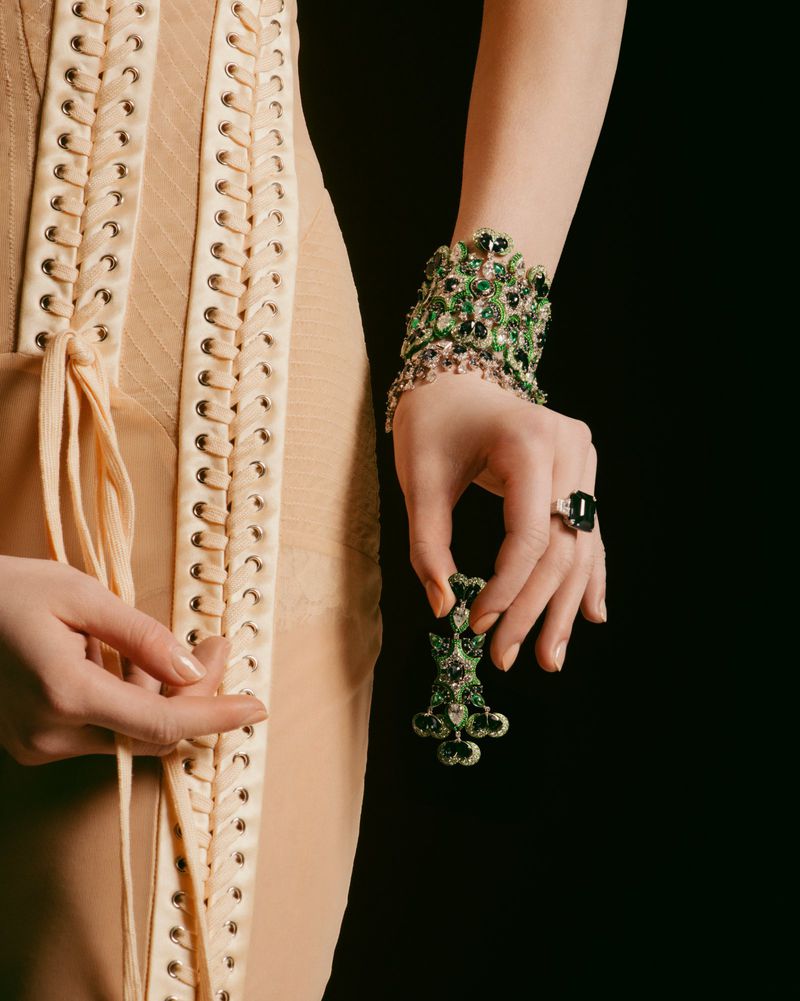 Model showcases green gemstone jewelry, including a large cuff bracelet and a ring, with a beige dress on a dark background.