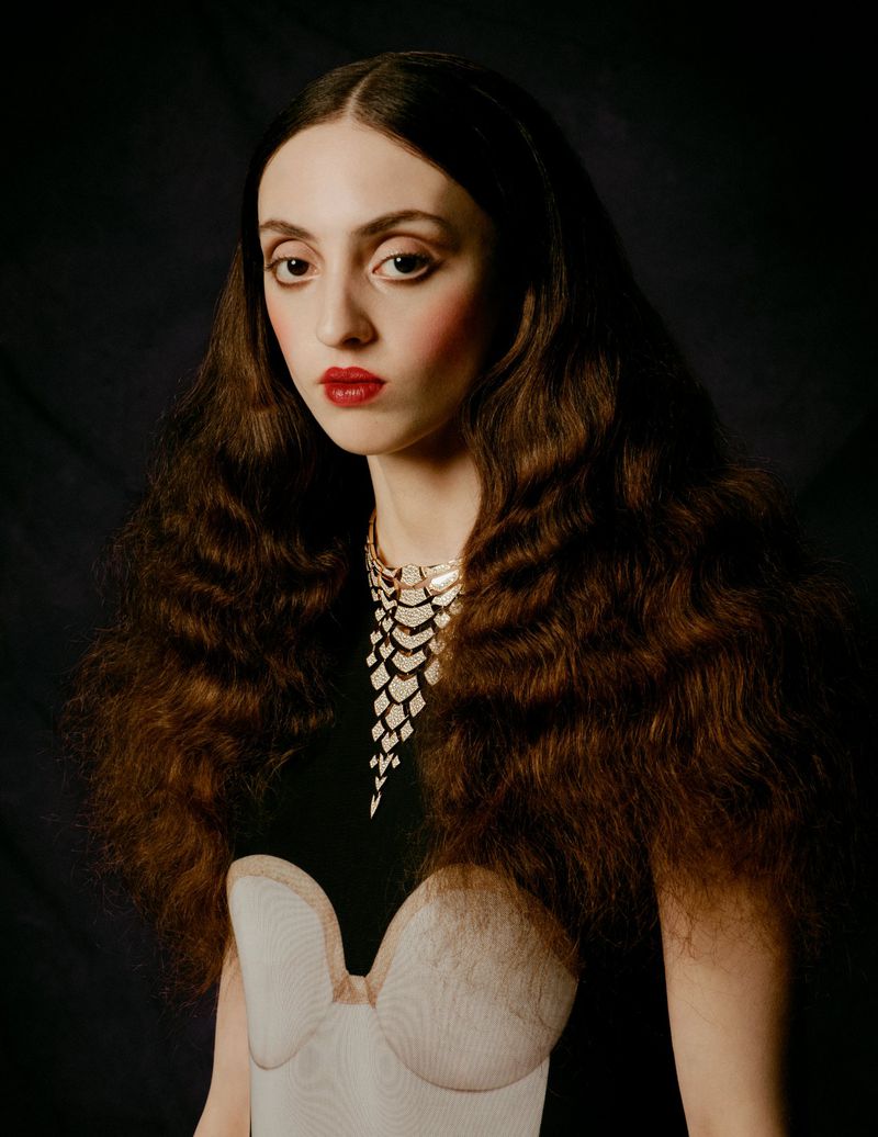 Portrait of a woman with long brown wavy hair, wearing a decorative gold and silver necklace, red lipstick, and a low-cut dress against a dark background.