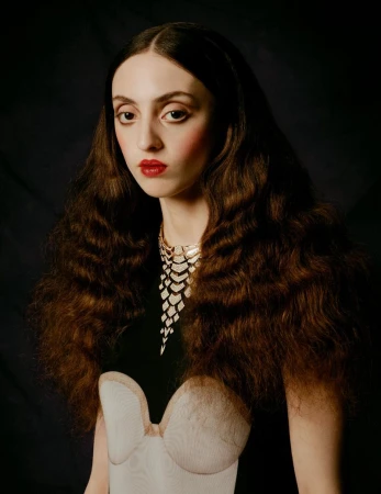 Portrait of a woman with long brown wavy hair, wearing a decorative gold and silver necklace, red lipstick, and a low-cut dress against a dark background.