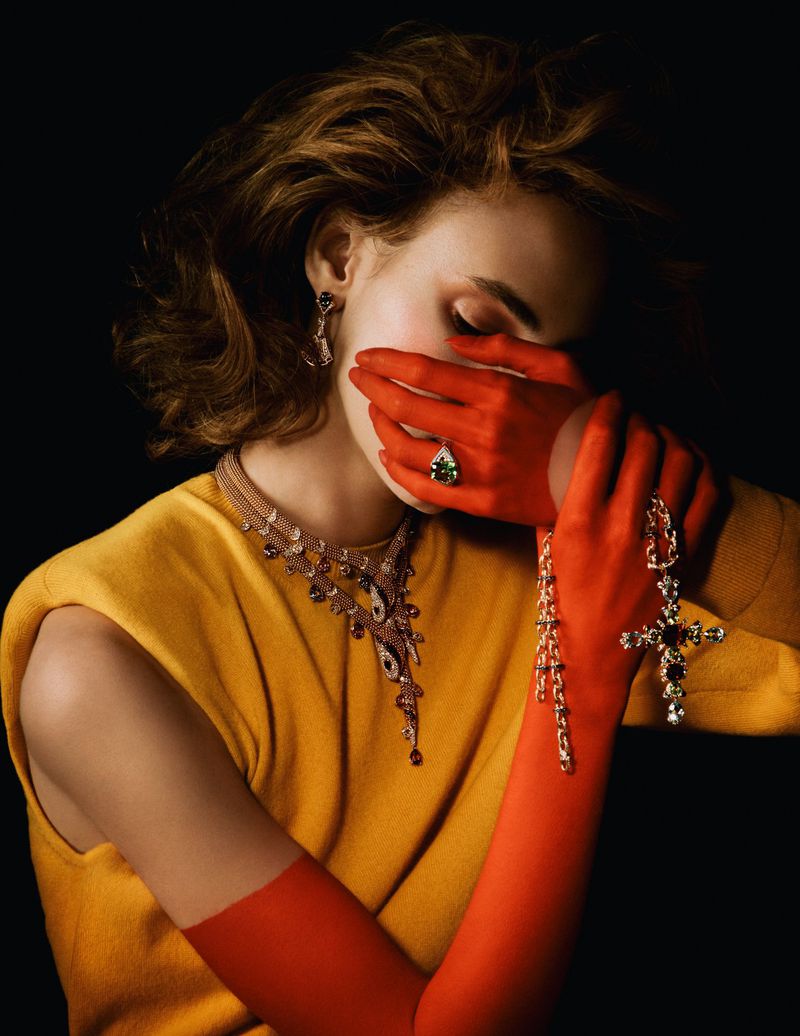 Indoor portrait of a blonde woman with red gloved hands covering her face, wearing a yellow sweater and an assortment of jewelry.