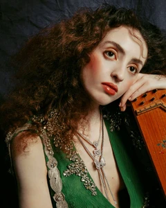 Portrait of a woman with curly hair wearing a green embellished dress and layered butterfly necklaces, leaning on a wooden instrument