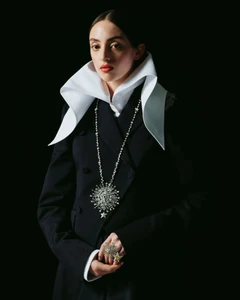 Model in dark double-breasted coat with large white collar and statement star pendant against black background