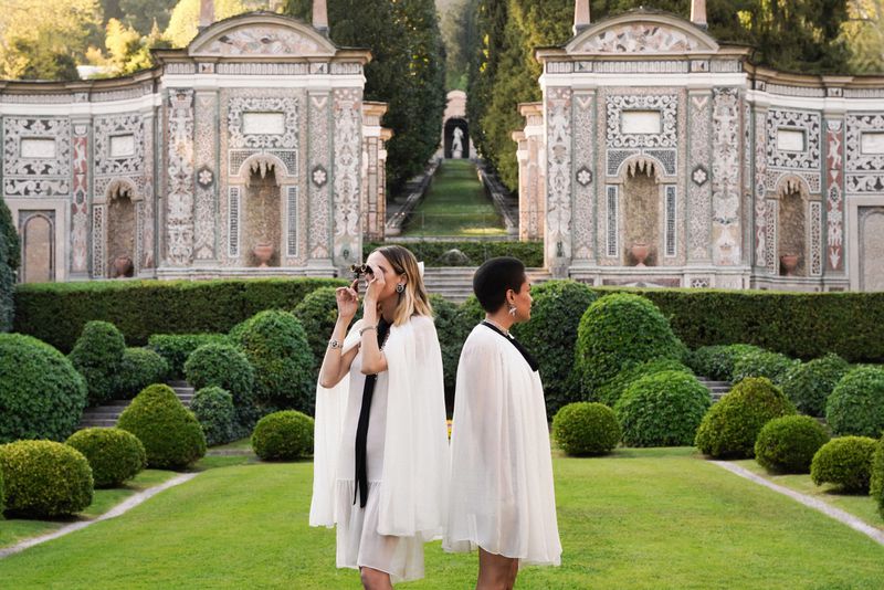 Two women in white dresses with capes stand in a formal garden with manicured bushes and ornate architecture, one using binoculars.