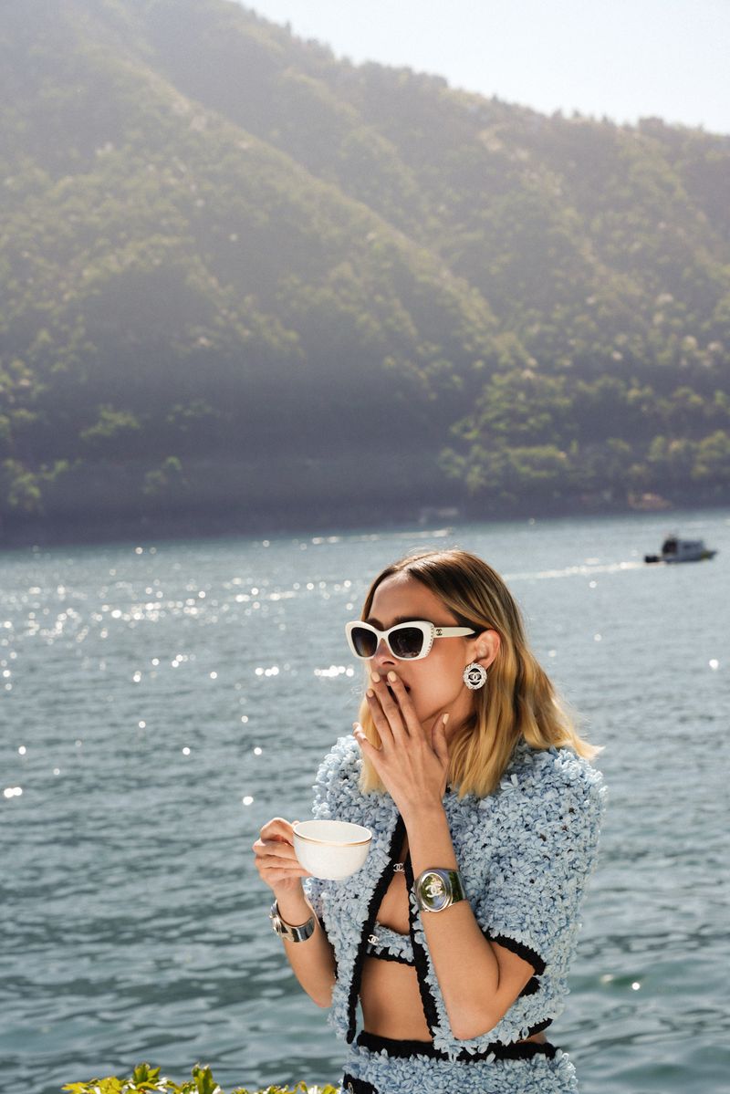 An adult female in a blue two-piece outfit and white sunglasses holds a teacup by a glimmering lake with mountains in the background.