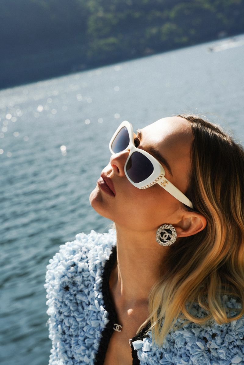Woman with sunglasses and Chanel jewelry poses outside near a body of water with a mountain in the background on a sunny day.