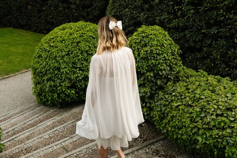 A woman with blonde hair, wearing a white dress, walks down stone stairs in a garden surrounded by green bushes in an outdoor setting.