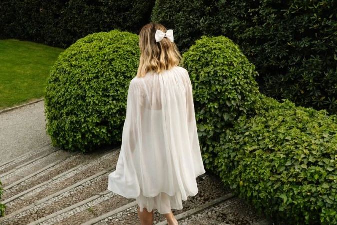A woman with blonde hair, wearing a white dress, walks down stone stairs in a garden surrounded by green bushes in an outdoor setting.