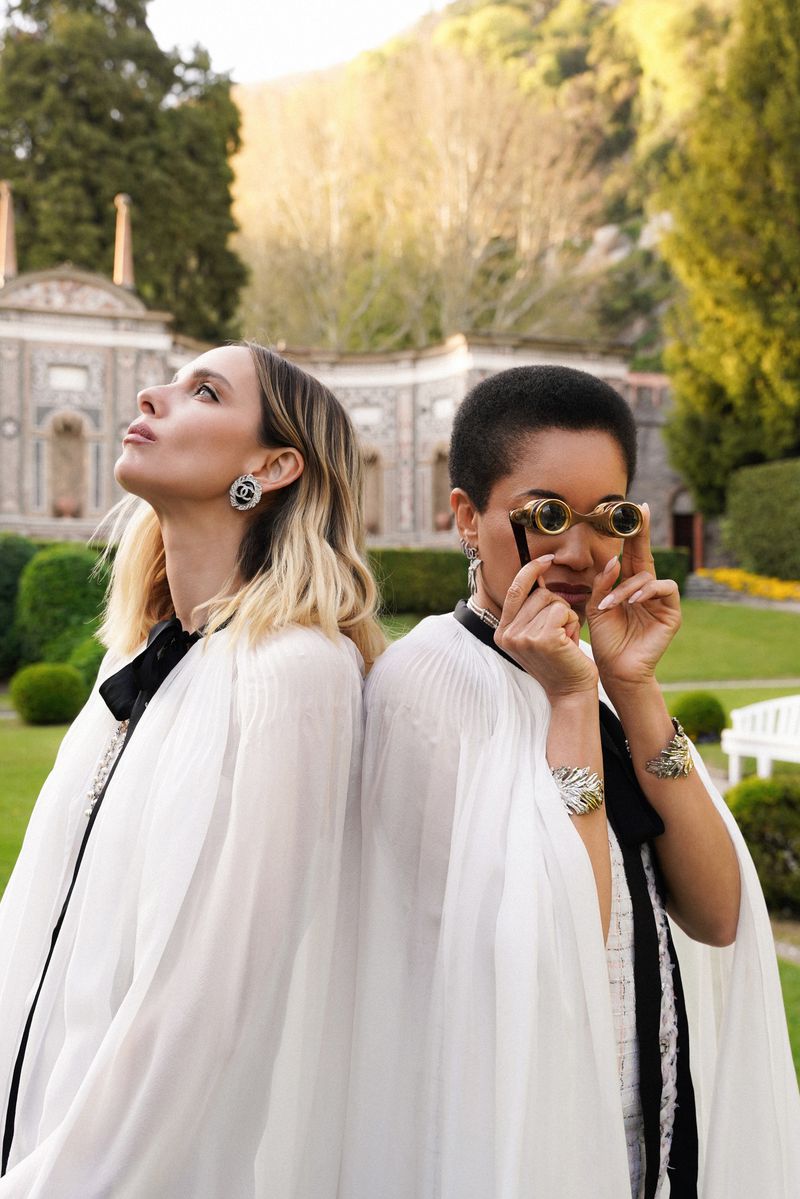 Two women stand back-to-back in a garden with vintage opera glasses, capes, and jewelry, with green trees and an ornate building behind them.