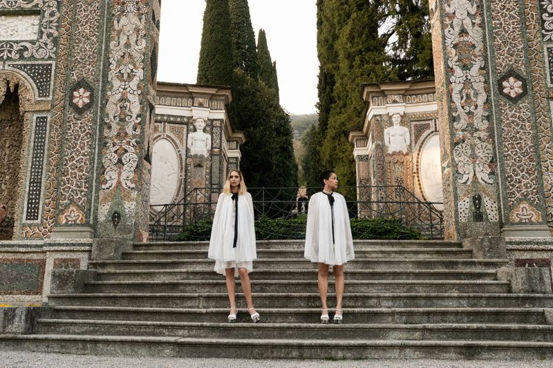 Two women stand on stone stairs in matching white dresses with black ties in front of an ornate building on a cloudy day.