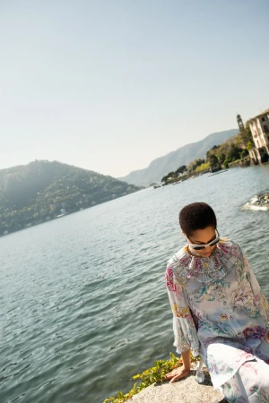 Woman in floral dress seated on stone ledge overlooking lake, mountains in distance, clear blue sky and water, outdoor fashion shot.