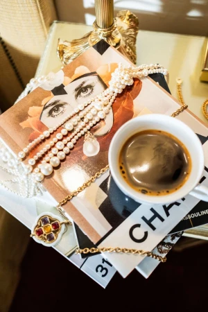 Still life featuring pearl necklaces, a cup of coffee, and fashion magazines on a table with golden lamp and decorative chair.