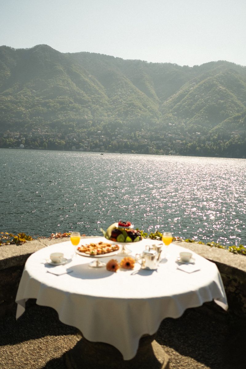 Outdoor breakfast on a round table with a white tablecloth, featuring pastries, fruit, juice, and a lake and mountain backdrop.
