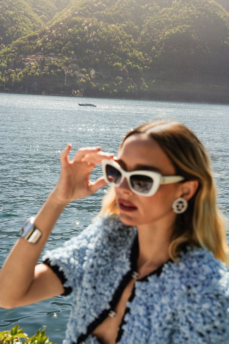 Woman in sunglasses near Lake Como, Italy. She is wearing a light blue jacket and silver bracelet, with mountains in the background.