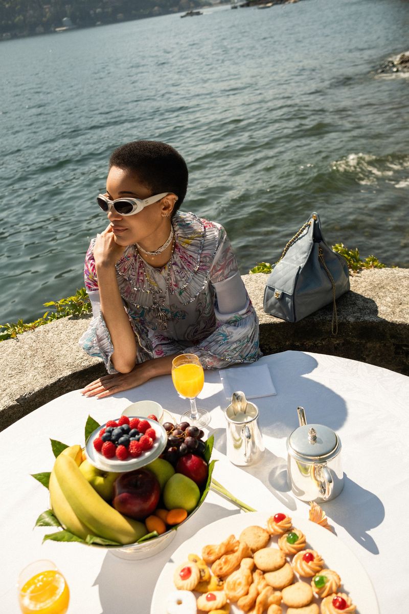 Elegant woman with sunglasses by Lake Como enjoys fruit, cookies, and juice on a white tablecloth with a blue water background.