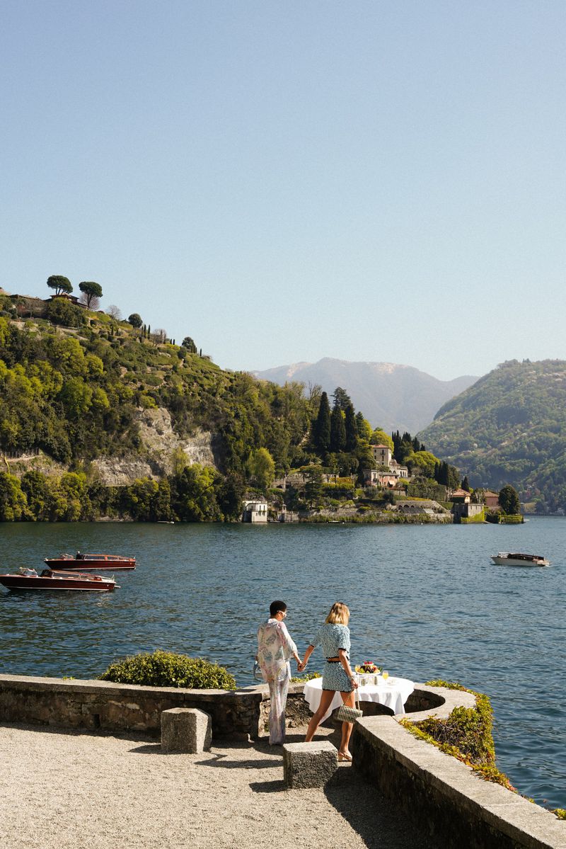 A couple walks along a stone terrace overlooking a lake with boats and mountains in the background on a sunny day in Lake Como.