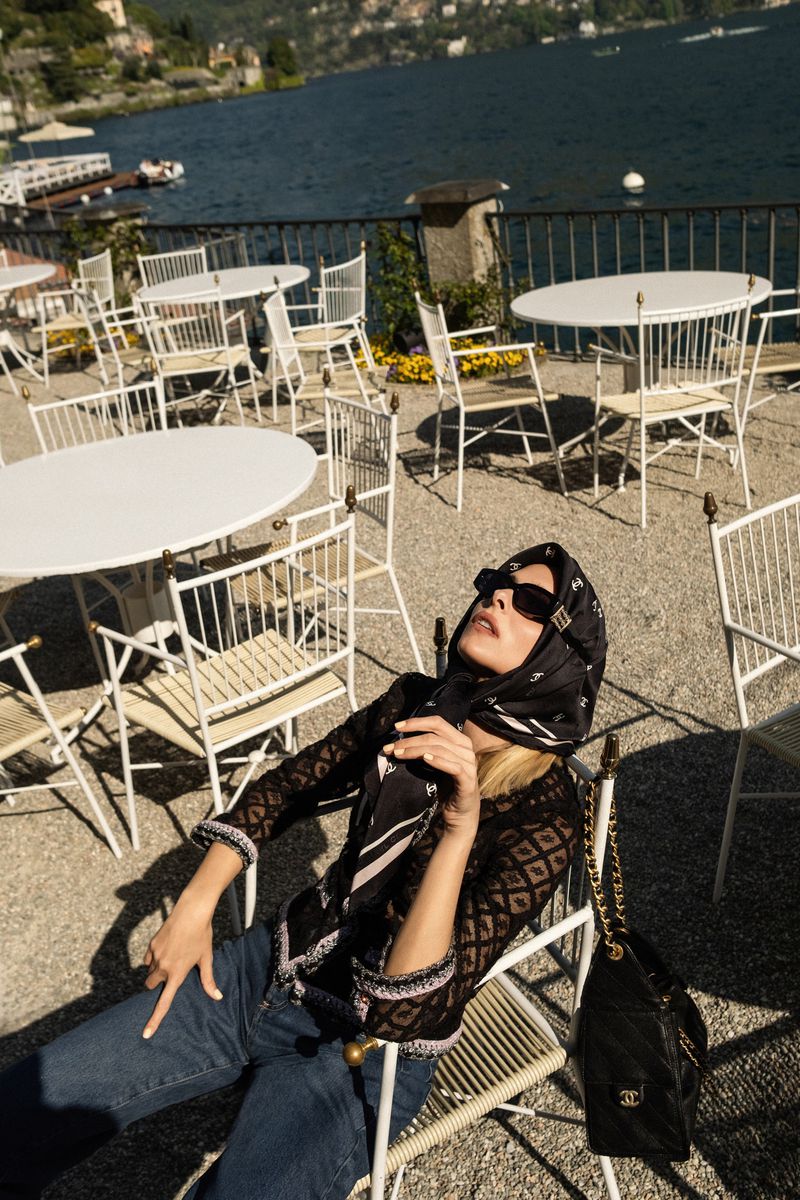Woman in headscarf and sunglasses reclines in chair at outdoor cafe with lake and mountains in the background on a sunny day.