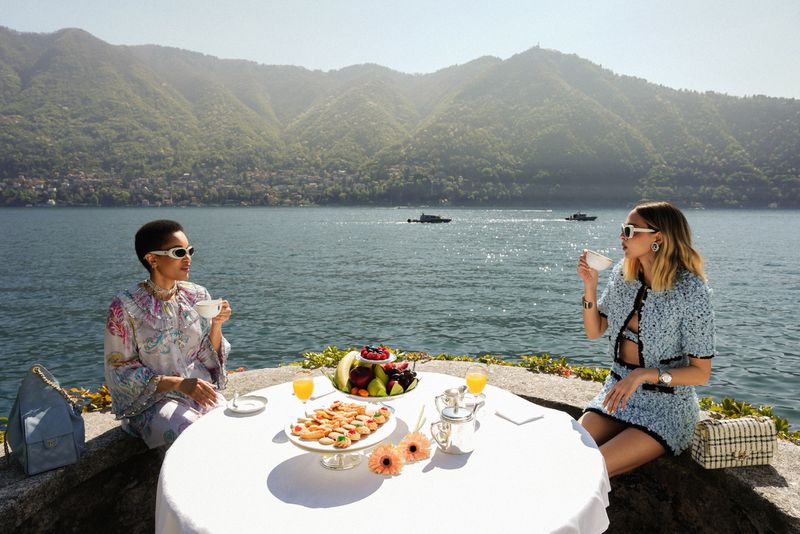 Two women enjoy tea on a stone terrace with lake and mountain views. Table with tablecloth, fruit, flowers, drinks, and handbags.