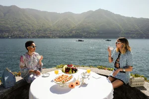 Two women having tea at an outdoor table overlooking a large lake and mountains, photographed by Alberto Alicata for L'Officiel Italia.