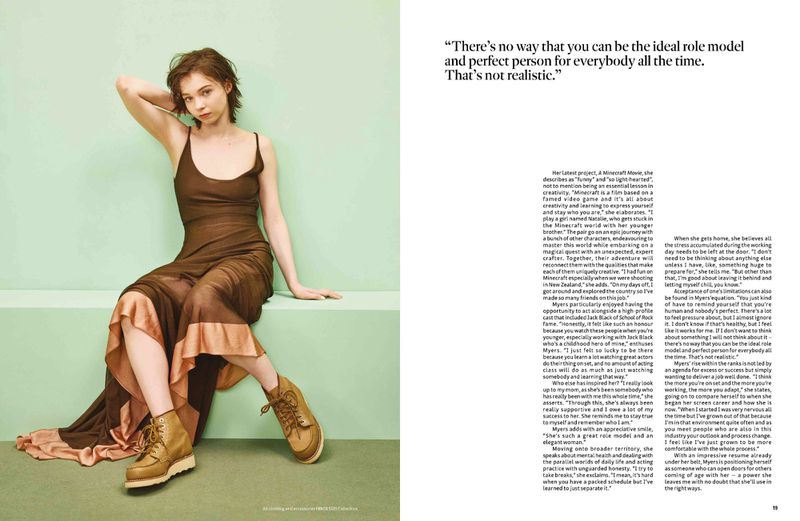 A young adult woman with a light complexion sits cross-legged on a bench wearing a sheer brown dress and lace-up boots in a studio setting.