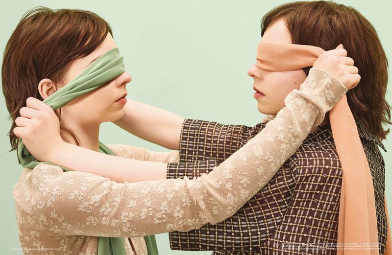 Two women facing each other, one tying a peach-colored blindfold on the other's eyes. The other woman has a similar green blindfold.