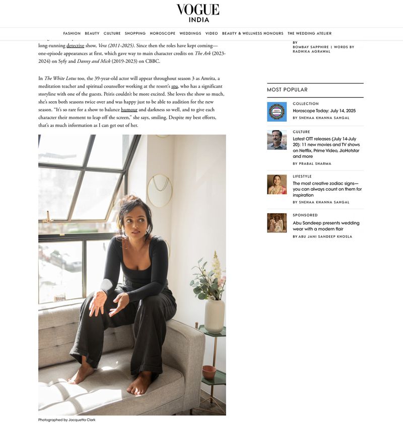 Indoor portrait of a South Asian woman in casual black top and pants sitting on a grey couch near a window with natural light.