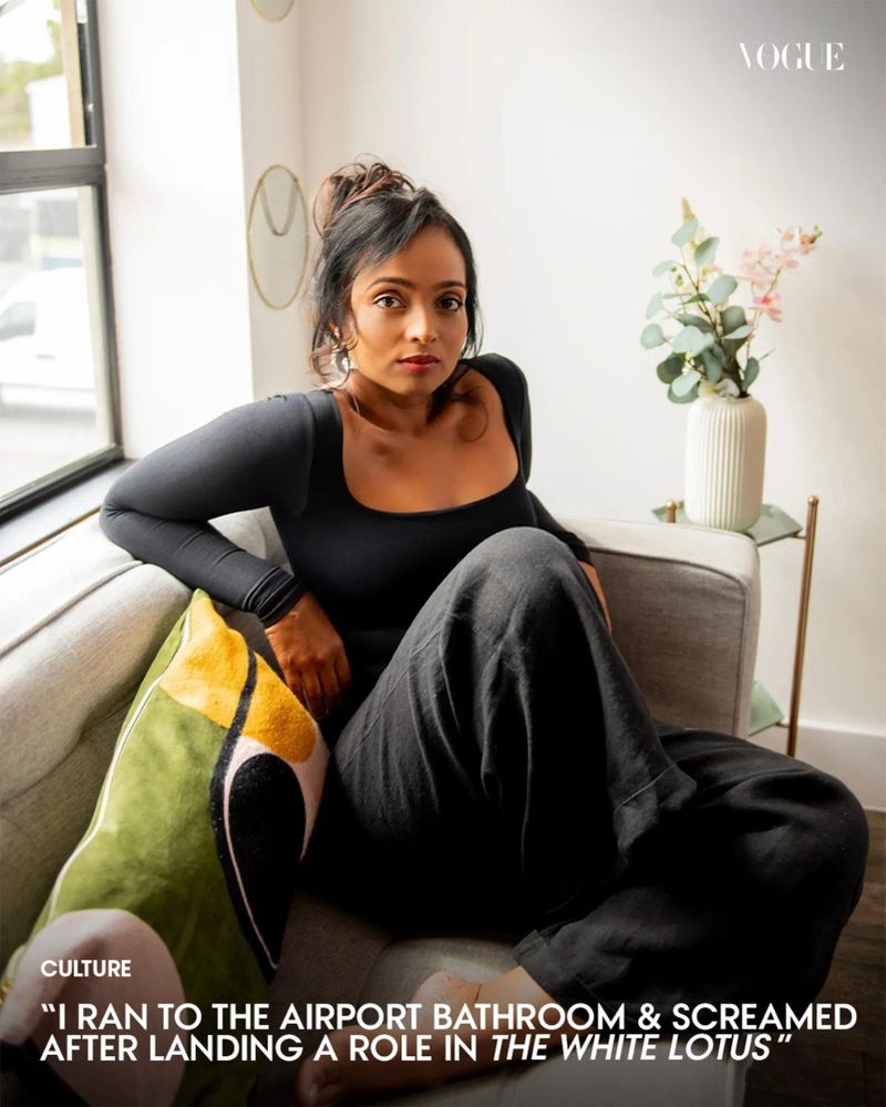 Young woman poses casually on a gray sofa in a brightly lit interior setting, complemented by a decorative pillow and vase of flowers.