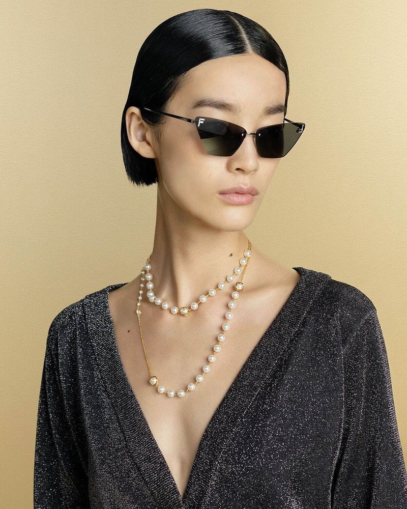 Portrait of an asian woman wearing sunglasses and a pearl necklace against a gold background in a studio.