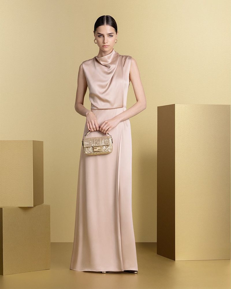 Model in a silk cowl-neck dress holding a small metallic handbag against a warm neutral studio set.