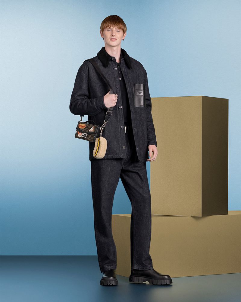 Full-length shot of a redhead male model in dark denim jacket and jeans, holding a small bag, in front of a blue and beige background.