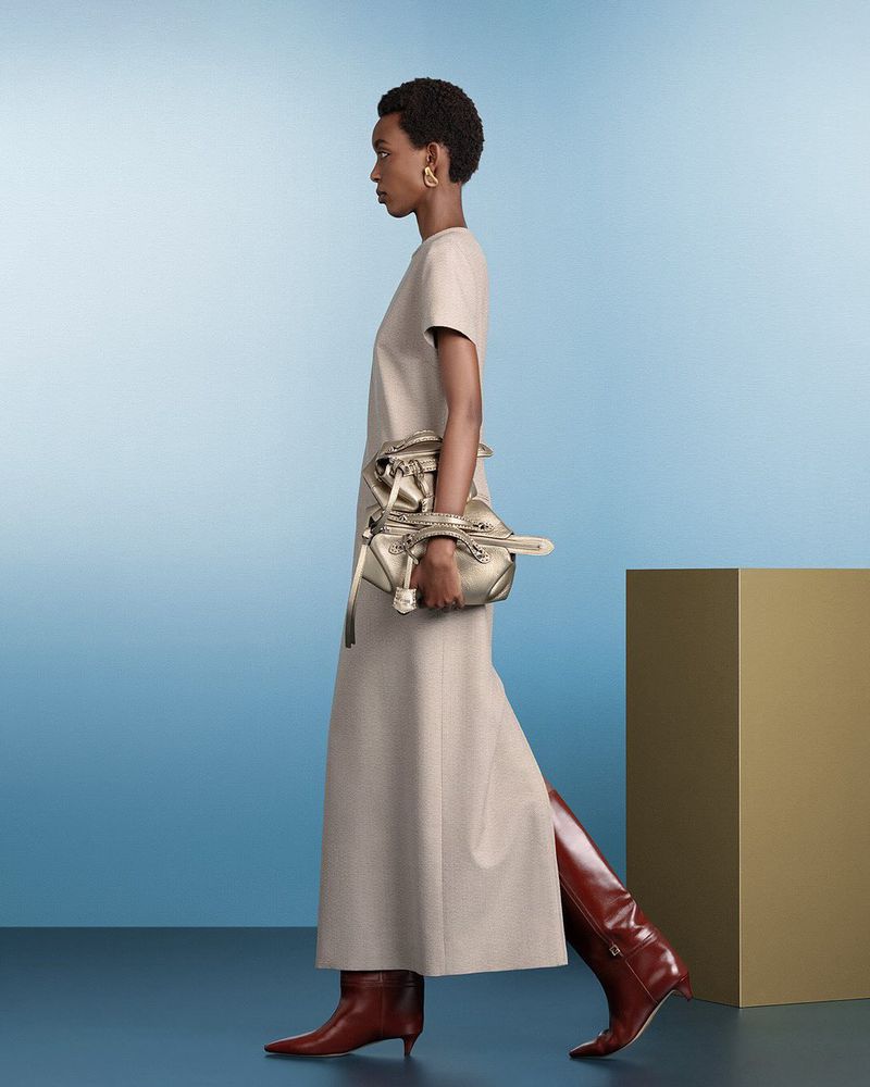 Fashion shoot featuring a woman walking in profile, wearing a dress and tall boots, carrying a metallic handbag against a studio backdrop.