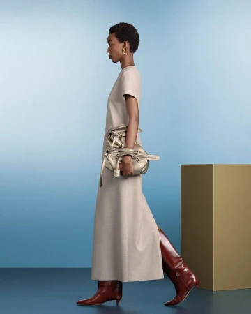 Fashion shoot featuring a woman walking in profile, wearing a dress and tall boots, carrying a metallic handbag against a studio backdrop.