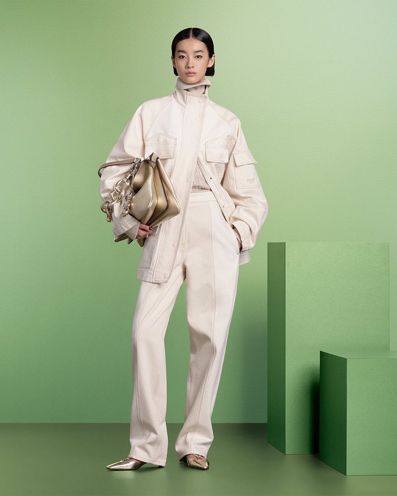 Studio shot of a woman in a cream-colored suit holding a gold handbag against a green backdrop with geometric blocks. Fashion editorial.