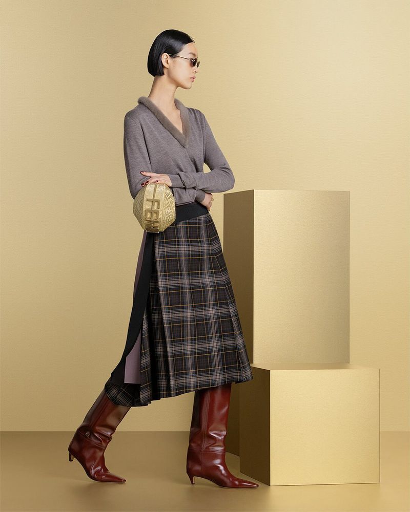 Fashionable woman in long-sleeve top, plaid skirt, and leather boots poses next to gold block, carrying a gold textured handbag.