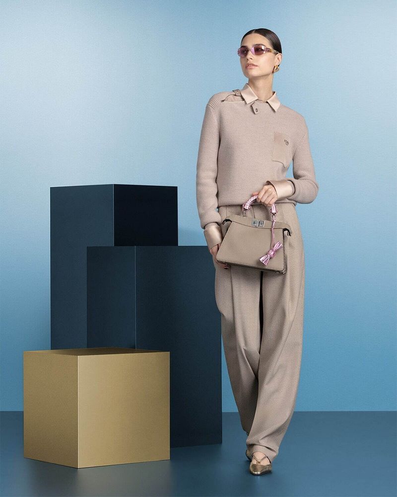 A stylish adult female model poses in a beige outfit with a handbag on a blue background with dark geometric cubes for a fashion editorial.