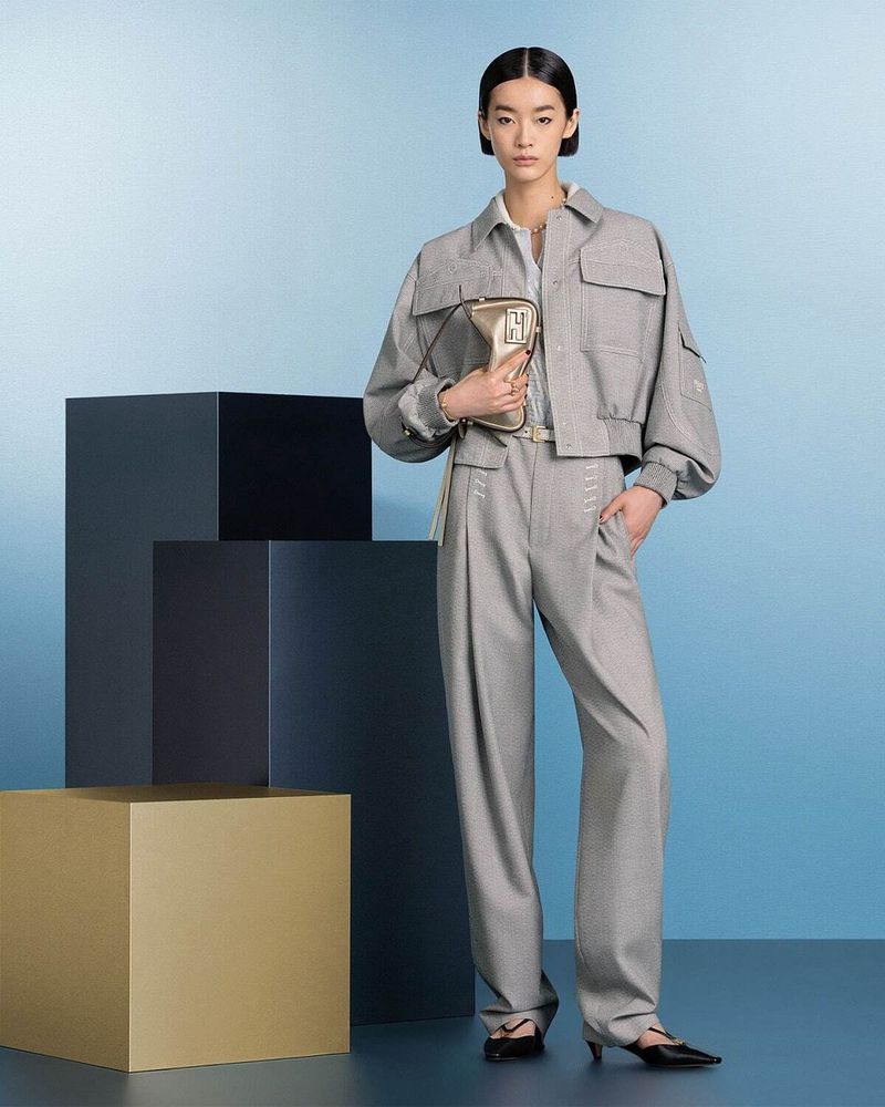Fashion model posing with handbag in grey patterned jacket and trousers on a light blue background with stacked cube props.