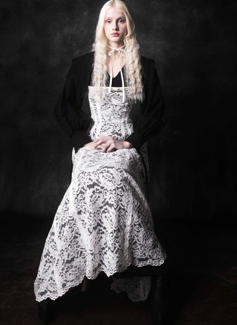 A young albino woman with long, pale hair poses seated in a white lace dress with a black jacket against a dark, textured backdrop.