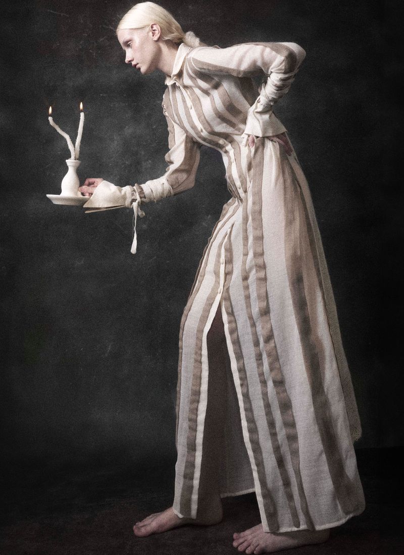Elegant woman in striped dress holding candles in a dark, moody setting. Striking pose, neutral tones, and unique candle holder on display.
