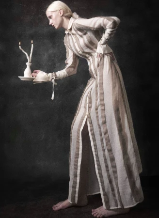 Elegant woman in striped dress holding candles in a dark, moody setting. Striking pose, neutral tones, and unique candle holder on display.