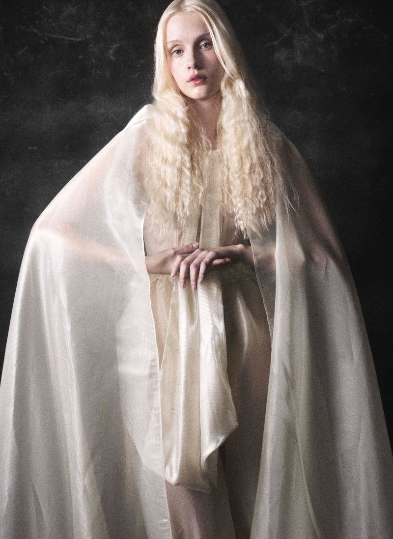 A blonde model with long wavy hair wears a sheer ivory cape draped over her shoulders against a dark background.