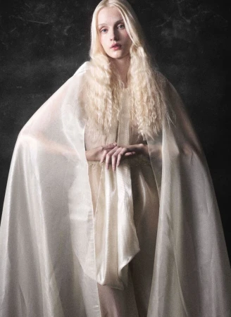A blonde model with long wavy hair wears a sheer ivory cape draped over her shoulders against a dark background.