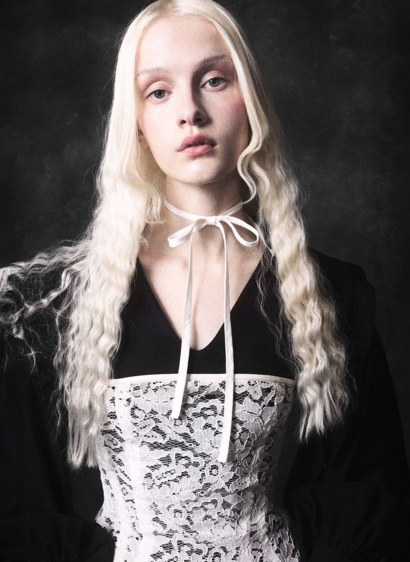 Portrait of a fair-skinned young woman with pale blonde wavy hair, wearing a white choker and lace top against a dark backdrop.