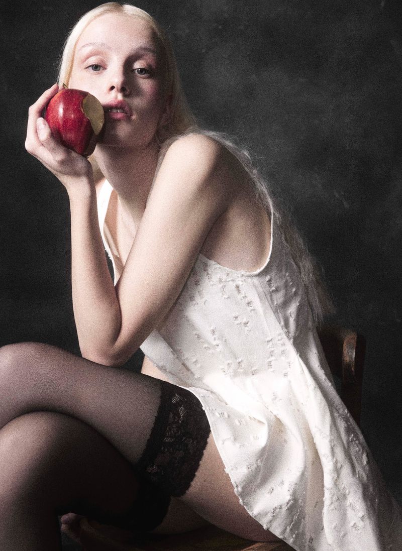 Blonde woman in negligee seated holding a red apple with a bite taken, wearing black stockings against a dark neutral background.
