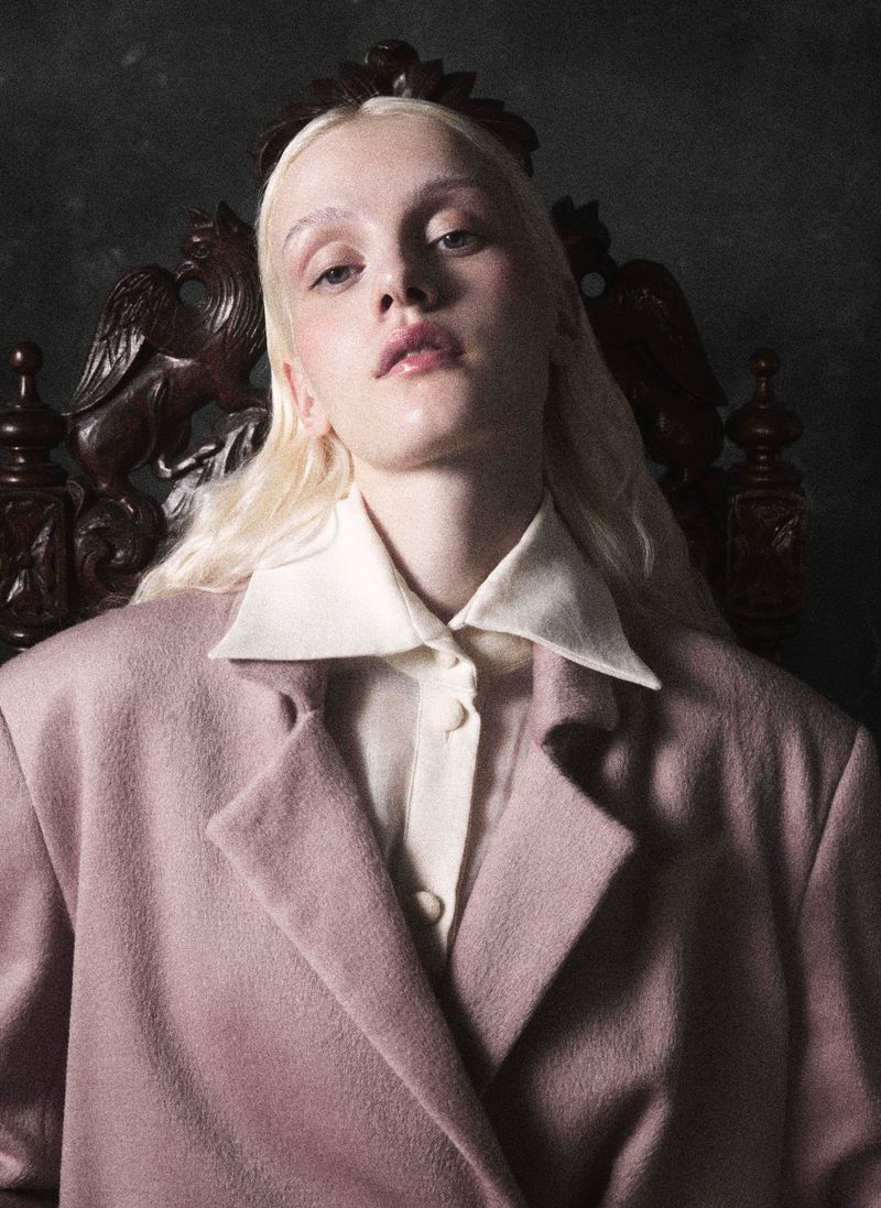 Portrait of an adult female with fair hair and wearing a blazer and collared shirt, sitting on an ornate chair in studio setting.