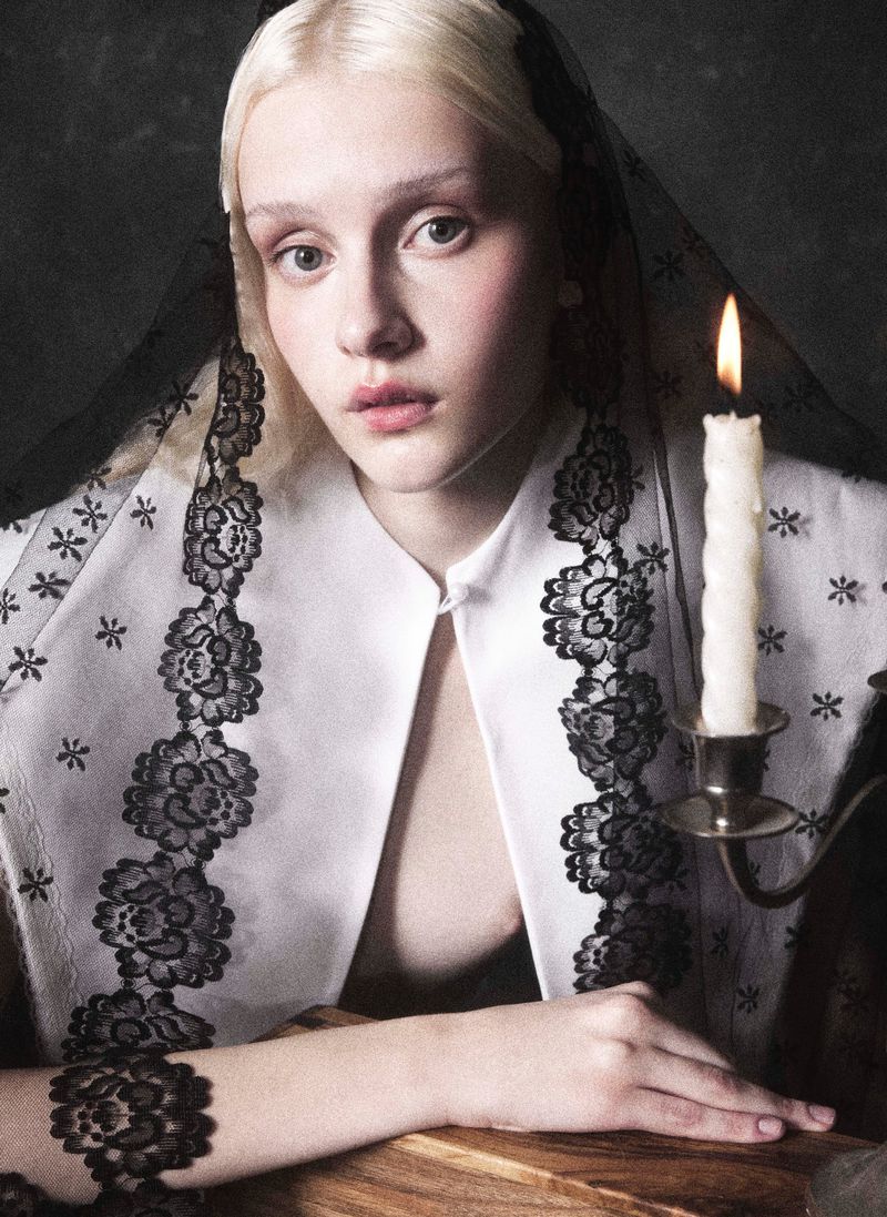 Portrait of a fair-skinned, fair-haired young adult woman wearing a lace-trimmed shawl, posing indoors with a lit candle in a holder.